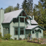 Another angle of a green renovated farmhouse amongst the trees, with a stack of fireood, raised bed and lawn in front.